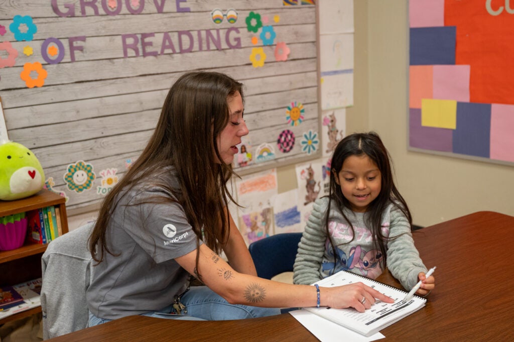 A woman sits next to a young student, pointing at a book they are reading together.