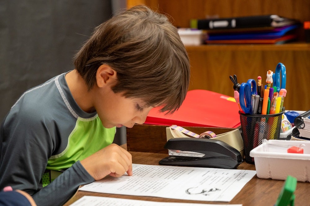 A student leans over a sheet of paper with a story printed on it, reading it and pointing to the words with his finger. 